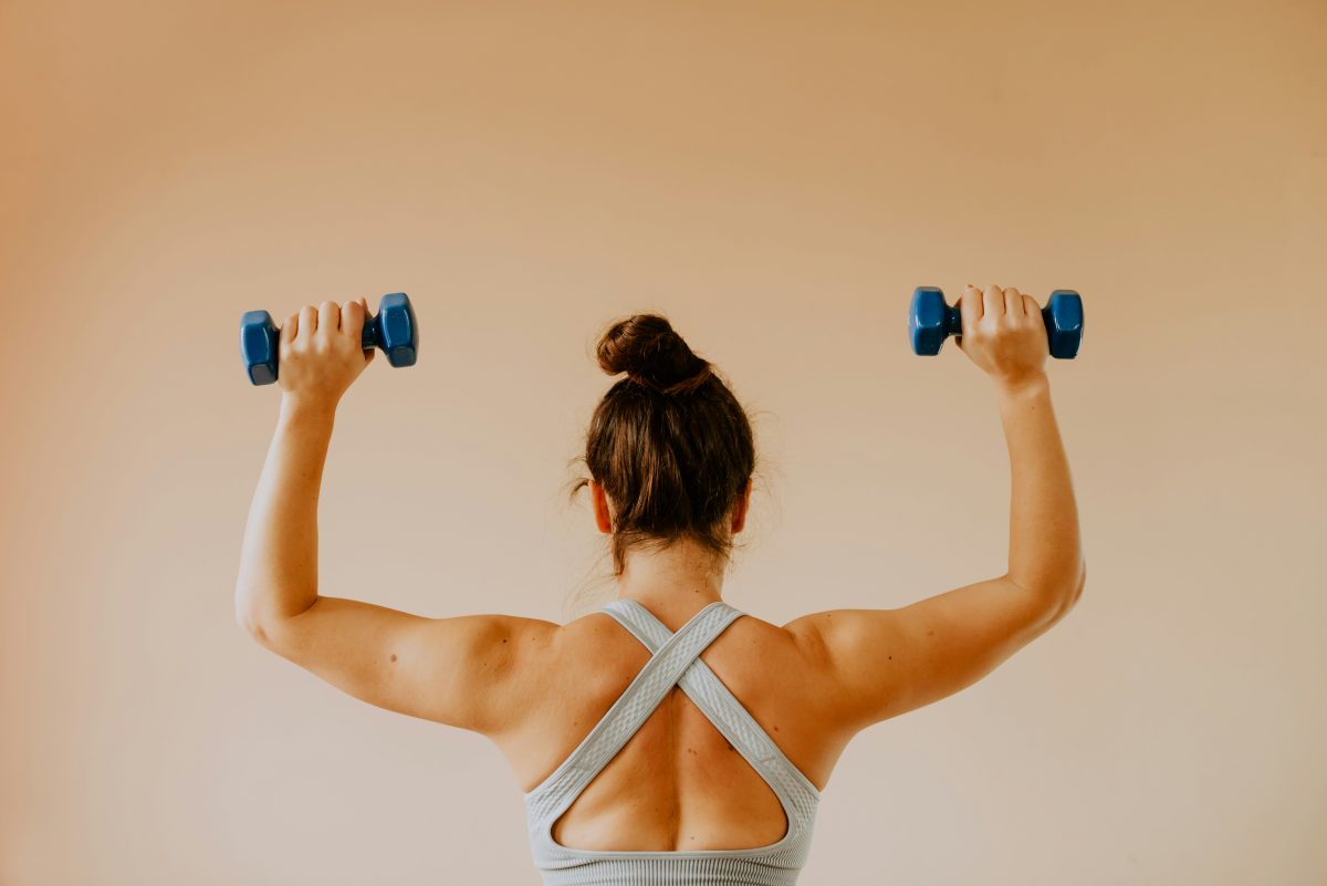 a young woman raises dumbbells with both arms, showing her toned shoulders and back.
