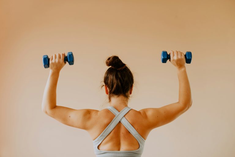 a young woman raises dumbbells with both arms, showing her toned shoulders and back.
